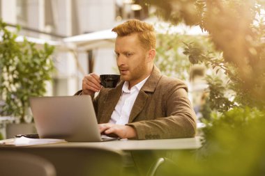 Konsantre olmuş yakışıklı adam laptopunda haber okurken kahvesinin tadını çıkarıyor.