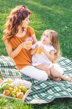 Kız ve anne parkta battaniyenin üzerinde oturmuş plastik bardaklardan portakal suyu içiyorlar.