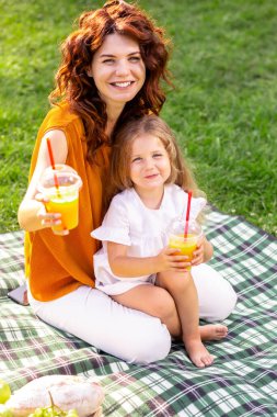 Anne ve kızı parkta battaniyenin üzerinde portakal suyu ile plastik bardaklar gösteriyorlar.