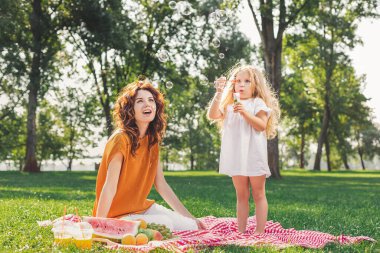 Annesi parkta piknik battaniyesinde otururken küçük kız köpük üflüyor.