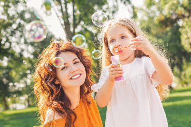 Anne, kızı parkta sabun köpüğü üflerken kameraya bakıyor.