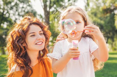 Parkta annesiyle sabun köpüğü üfleyen kızın portresi.