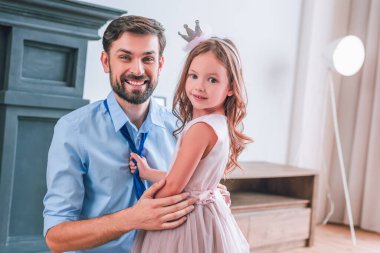 Küçük kız babasına kravat bağlıyor ve evdeki kameraya bakıyor.