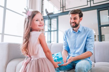 Oturma odasındaki kanepede oturan babasına hediye paketi sunan kızın portresi.