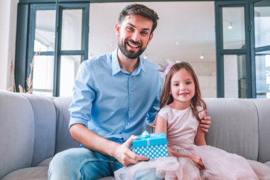 Hediye kutusuyla kanepede oturan ve kameraya bakan kız ve baba portresi.