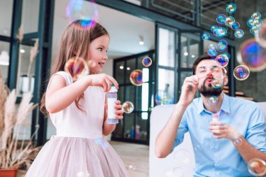 Baba ve kızının oturma odasında sabun köpüğü üflerken portresi.
