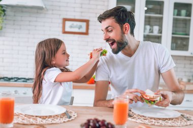 Kız, babamı sandviçiyle besliyor. Kahvaltıda mutfakta yiyor.