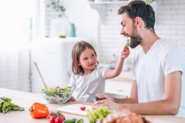 Kızı babasını beslerken mutfakta salata hazırlıyor.
