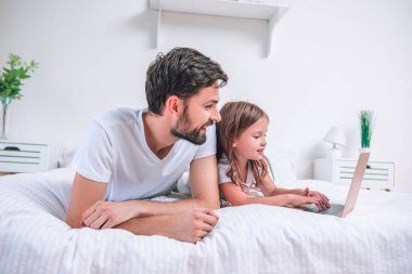 Baba ve kızı yatağın üzerinde dizüstü bilgisayar kullanıyorlar.