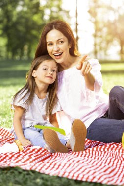 Piknik battaniyesinde otururken parkta kızına bir şey gösteren bir kadın.