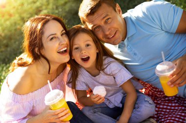 Baba, anne ve kız parkta piknikte kameraya bakıyorlar.