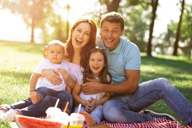 Ebeveynler parkta battaniyenin üzerinde oturup kameraya bakarken kızlarına ve oğullarına sarılıyorlar.