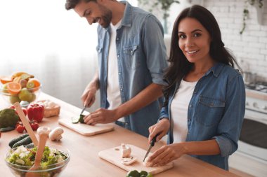 Mutfakta bir adamla sebze doğrarken kameraya bakan gülümseyen bir kadın.
