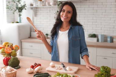 Esmer kadın yemek pişirirken spatula tutuyor ve mutfaktaki kameraya bakıyor.