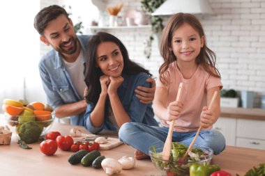 Anne ve baba mutfakta salata karıştırıyor ve kameraya gülümsüyor.