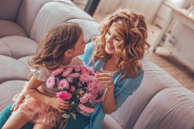 Kanepede oturan ve birbirlerine bakan bir buket çiçekle annelerinin kucağında oturan kızı.