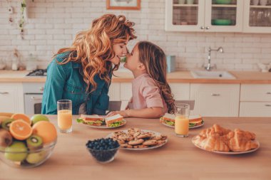 Mutfak masasında yemek yerken burnuna dokunan anne ve kızın yan görüntüsü.