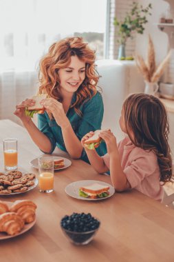 Anne ve kızı kahvaltıda sandviç yiyorlar ve mutfakta birbirlerine bakıyorlar.
