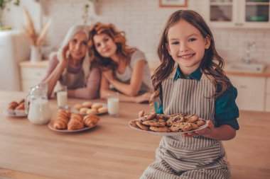Elinde bisküvi olan neşeli bir kız ve anne ve büyükannesinin mutfak masasında oturduğu kameraya bakıyor.