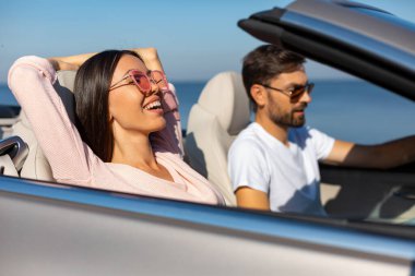 Genç bir kadın ve erkek güneşli bir günde üstü açık arabada ata biniyorlar.