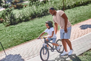 Düşünceli Afrikalı Amerikalı bir adam bisiklet sürerken çocuğunun dengesini korumasına yardım ediyor.
