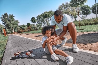 Yararlı bir Afro-Amerikalı baba, kaykaydan düştükten sonra çocuğunun hafif yaralandığını kontrol ediyor.