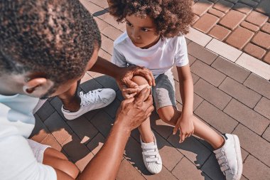 Yakışıklı Afro-Amerikalı bir çocuk babasına bakıyor. Kaldırımda otururken dizine bandaj yapıştırıyor.