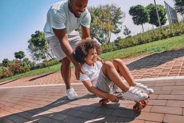 Neşeli bir Afro-Amerikalı çocuk kaykayın üstüne oturmuş ve babası onu itmiş.