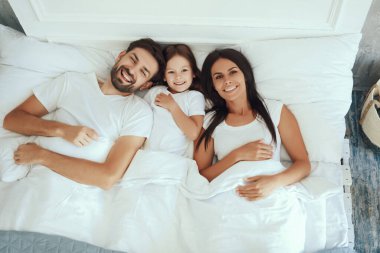 Annesi ve babasının arasında beyaz gömlekler içinde yatarken gülümseyen mutlu küçük kız.