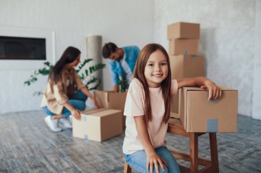 Sevimli küçük kız, ailesi arka plandaki eşyaları yerleştirirken heyecanlı ve kahverengi bir kutuya yaslanmış görünüyor.
