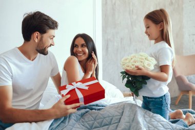 Koyu saçlı neşeli bir kadın yatakta oturmuş kocasına bakıyor ve küçük tatlı bir kızdan çiçekler alıyor.