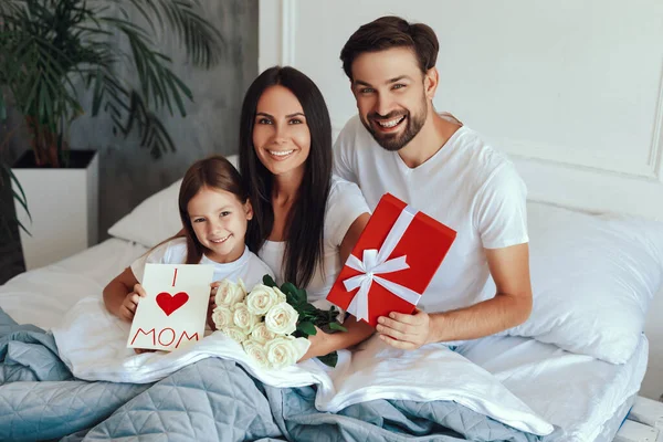 Merry little family smiling and staying under bed covers while holding mother day gifts and looking at the camera