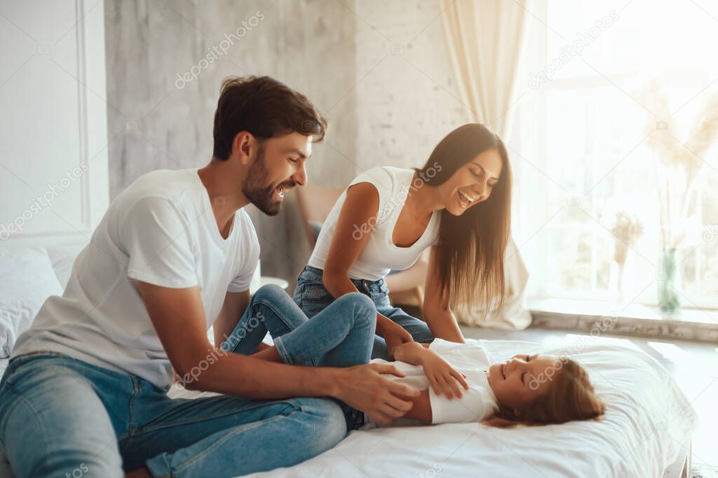 Merry young parents having fun with their little daughter while staying indoors and laying on a bed