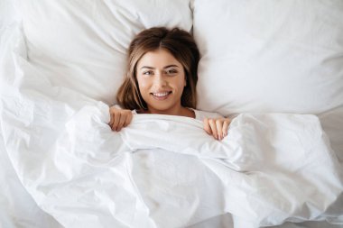 Positive young lady with a shiny smile looking at the camera while staying under the blanket in her bed clipart