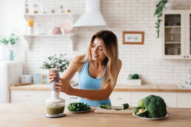 Blender 'da yeşil sebze kokteyli yapmak için çalışan heyecanlı genç bir kadın.