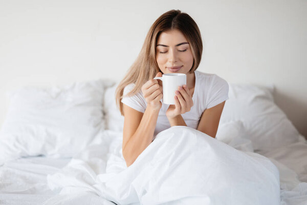 Blissful young lady closing her eyes and smelling the aroma of fresh coffee while chilling in her bed
