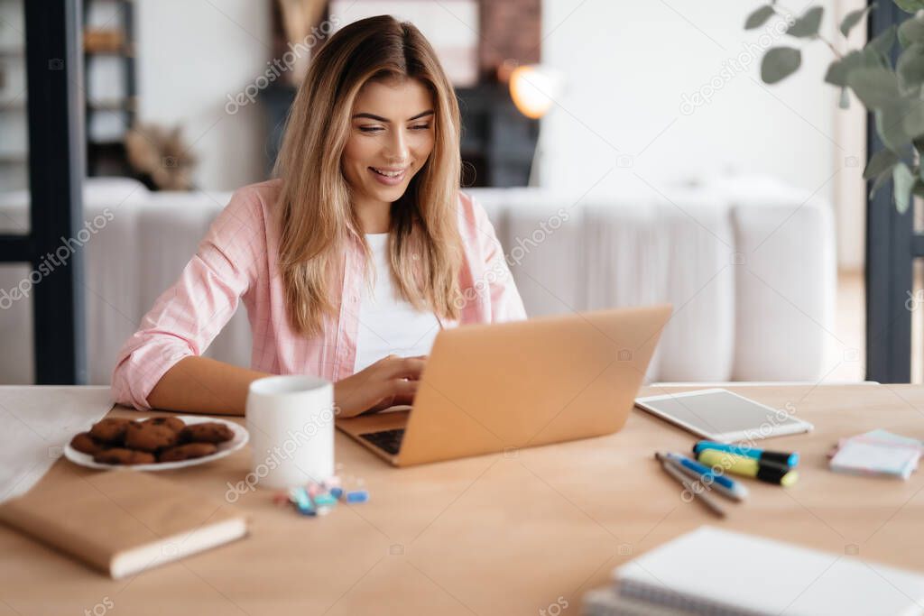 Joyous young lady smiling while sitting at working desk and looking at laptop screen