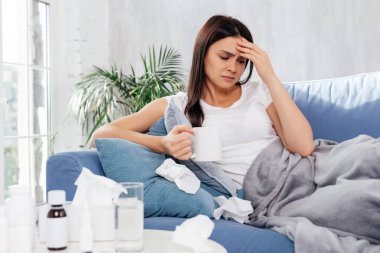 Kaşlarını çatmış bir kadın elinde bir bardak sıcak içecekle elini alnına koyuyor.