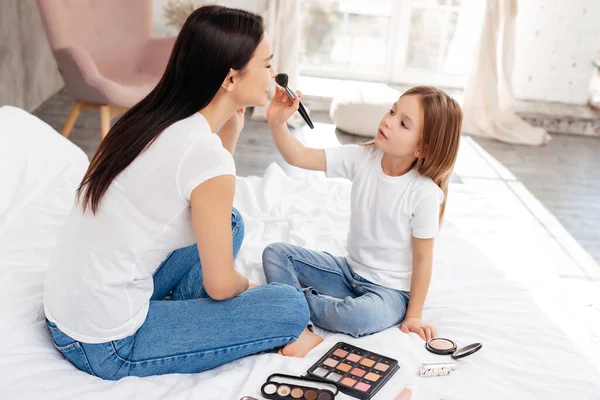 Focused child touching woman nose with a powder brush while sitting on white bed