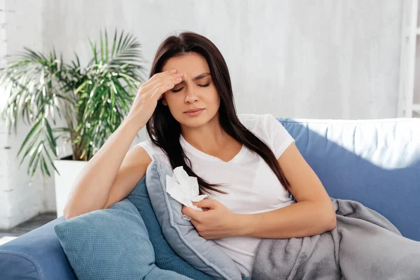 Worried female touching her forehead and keeping eyes closed while resting on sofa