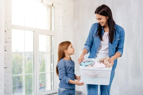 Neşeli anne yıkamak için giysi dolu bir konteynırı tutarken pozitif kızına bakıyor.