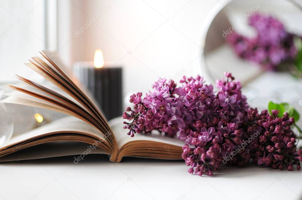 Cozy spring atmosphere at home with lilac, book and candle. Blooming time.