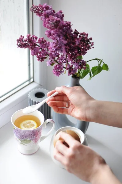 Vista superior da mão da mulher adicionando açúcar a uma xícara de chá. Flores de lilás no fundo — Fotografia de Stock