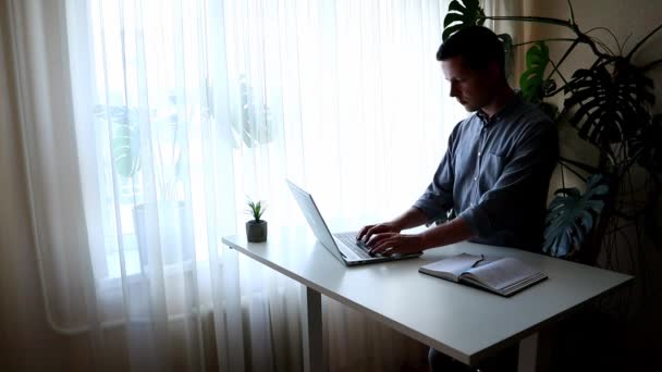 Jovem está de pé, trabalhando no laptop e escrevendo algo no caderno — Vídeo de Stock