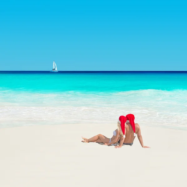 Couple in christmas hats at ocean beach with yacht — Stock Photo, Image