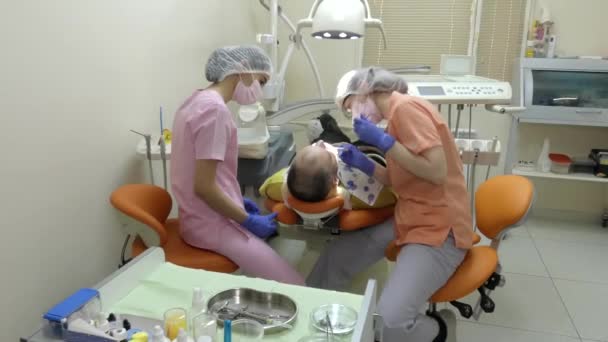 歯医者の所で医者は人の歯を治療する 近くで 若いアシスタントが彼女の仕事を見て — ストック動画