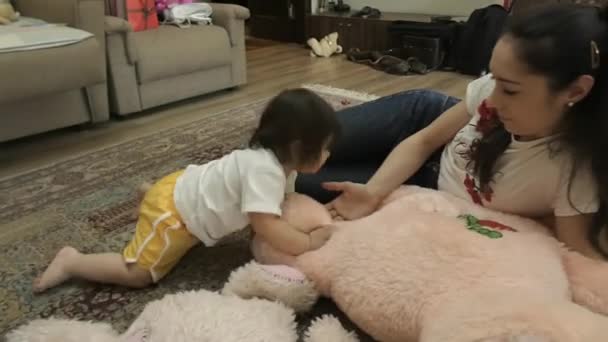 这孩子爱抚他的母亲 一个年轻的女人躺在房间里柔软的玩具上 不远处 她的孩子爬到地毯上 然后抱着她的母亲 — 图库视频影像