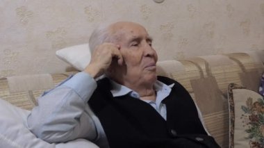 Yaşlı adam kanepede mutlu bir şekilde gülümsüyor, yastığa yaslanıyor. Başını yana çevirir, birine bakar..
