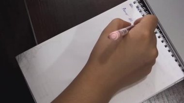 Bir kadının eli deftere bir şey yazıyor. Yanında bir dizüstü bilgisayar var. Yukarıdan görüntüle.