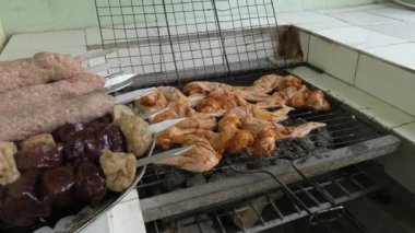 Bir el ızgaranın üzerine marine edilmiş tavuk kanadı koyar. Solda bir tabak çiğ et kebabı var..